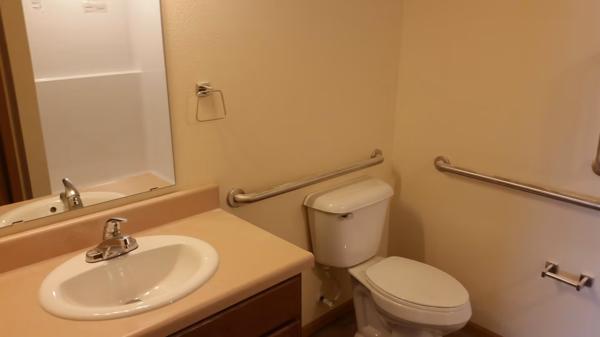 A bathroom with a white sink and faucet on a beige countertop, a large mirror above the sink, a white toilet with a closed lid, beige walls, and metal grab bars installed on the walls for support.