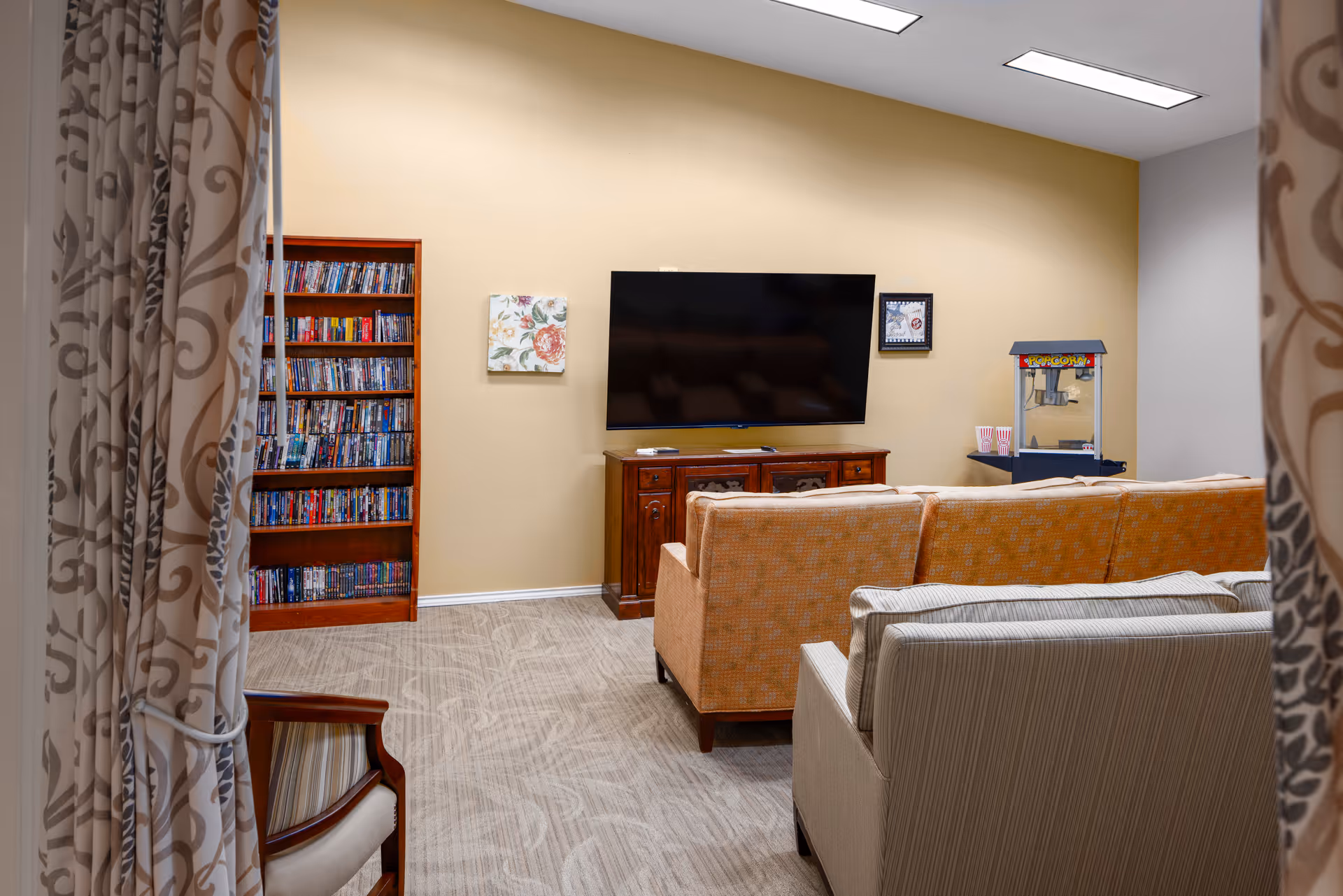 A cozy common room with sofas facing a wall-mounted TV, a bookshelf and a popcorn machine.