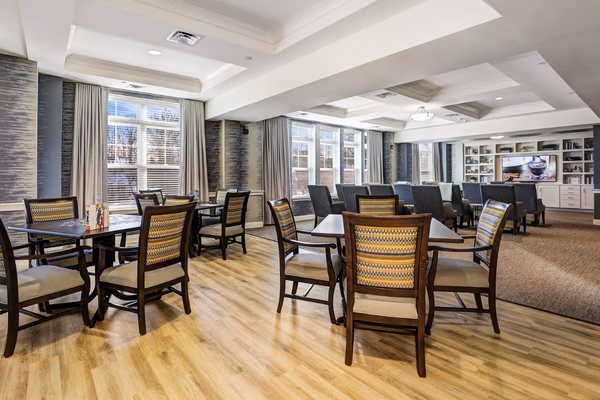A spacious and well-lit common area in a senior living facility with multiple wooden tables and chairs arranged on a light wood floor. Large windows with beige curtains allow natural light to fill the room. In the background, there is a seating area with armchairs facing a large flat-screen TV mounted on a wall with built-in shelves and cabinets.