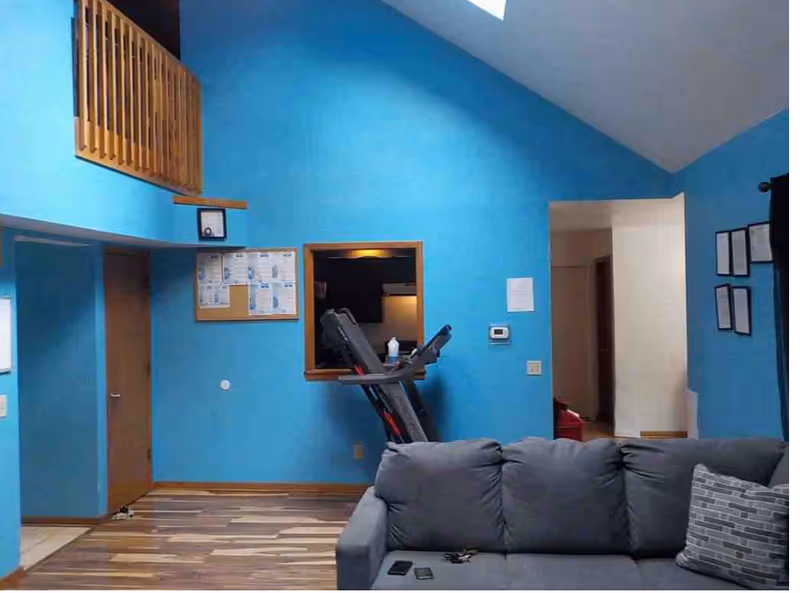 Common room with bright blue walls, a gray couch in the foreground, a treadmill against the wall, and bulletin board and doorways.