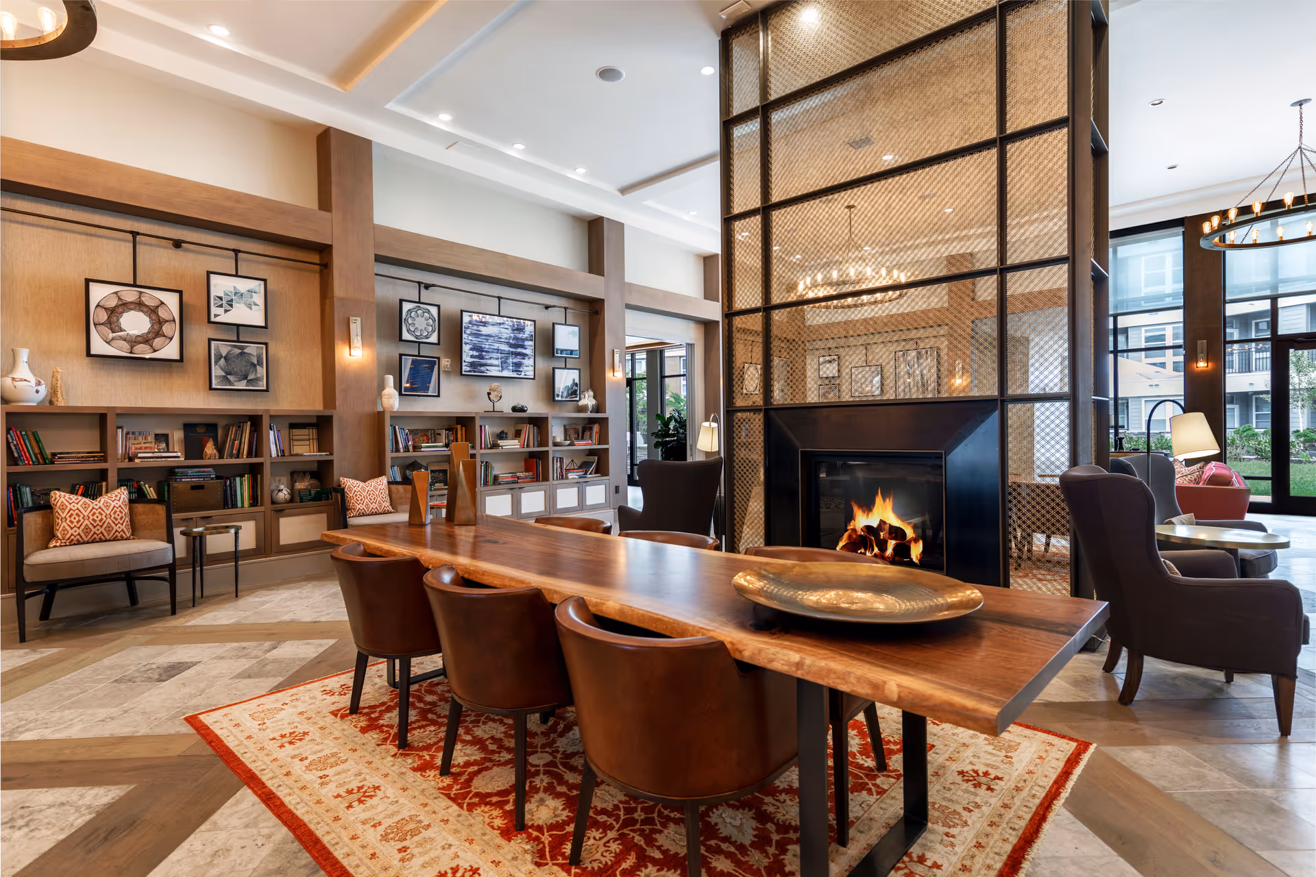 Spacious communal lounge with a long wooden table and leather chairs, a lit fireplace, bookshelves and additional seating by large windows.