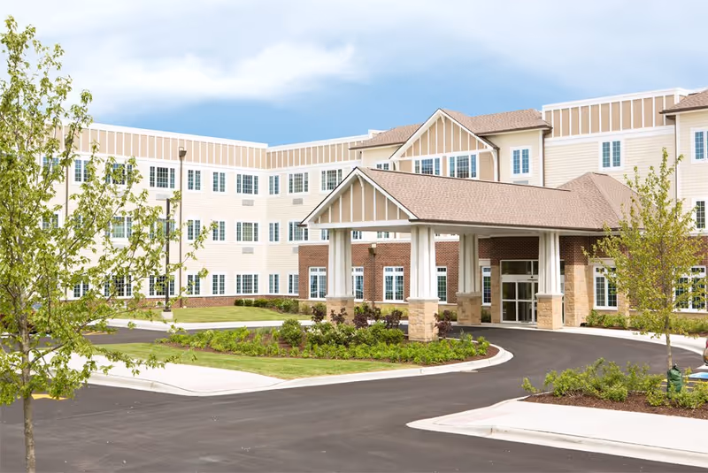 Exterior view of a large, multi-story senior living facility building with beige and white siding, numerous windows, and a covered entrance driveway surrounded by landscaped greenery and young trees under a partly cloudy sky.