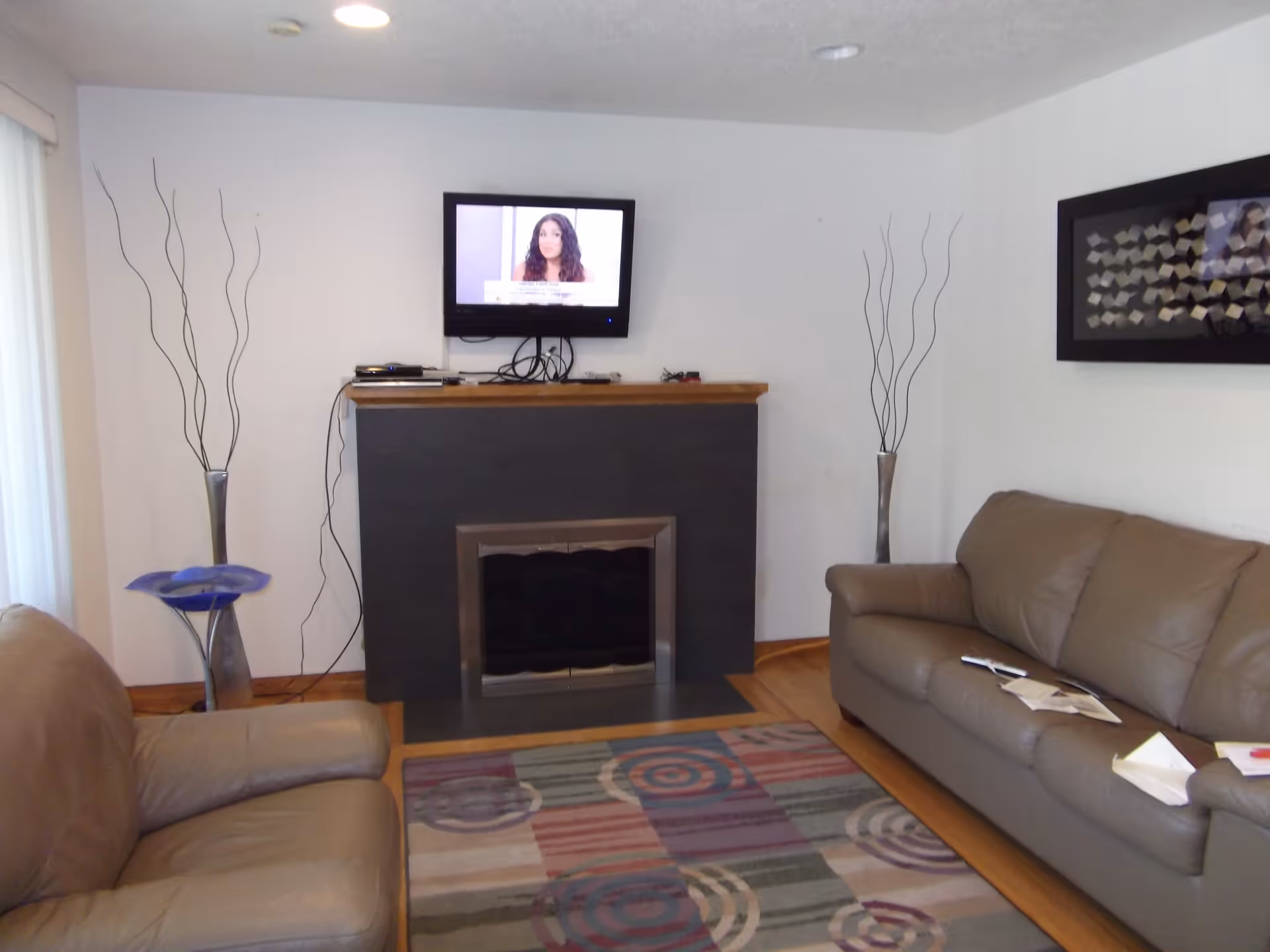 A living room with two brown leather sofas facing a fireplace with a mounted flat-screen TV above it. There are two tall decorative vases with thin branches on either side of the fireplace, a colorful patterned rug on the wooden floor, and a framed abstract artwork on the wall to the right.
