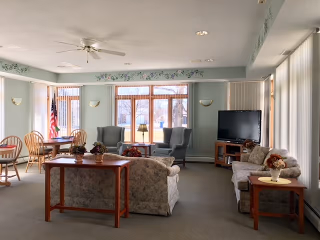 A spacious living room area with floral-patterned sofas and armchairs arranged around wooden tables. Large windows with vertical blinds allow natural light to fill the room. A flat-screen TV is placed on a wooden stand in the corner. There is a ceiling fan and wall-mounted lights, with an American flag visible near a small dining table with wooden chairs.