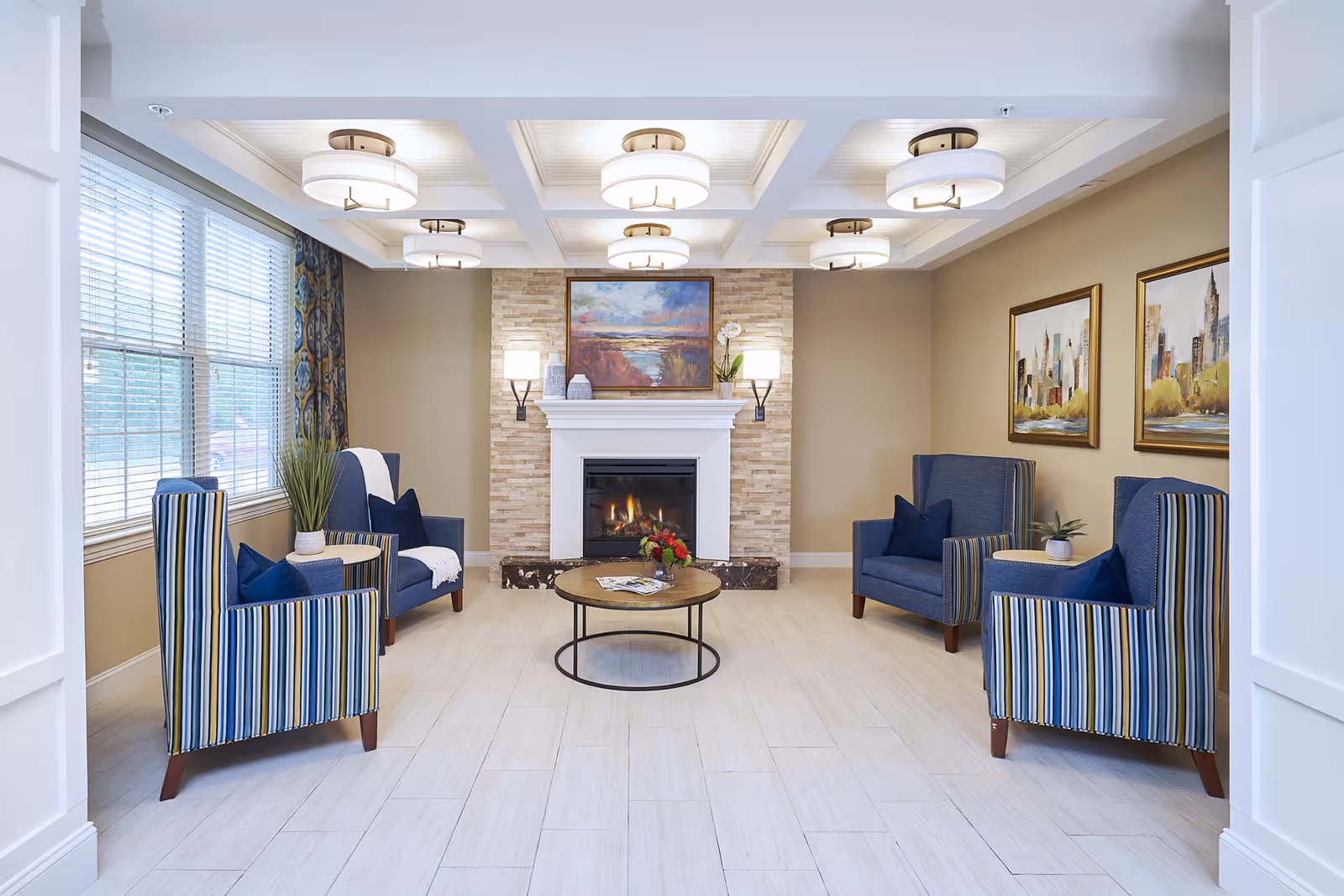 A bright and inviting living room with four blue armchairs arranged around a round coffee table with a flower arrangement. The room features a fireplace with a stone surround and a colorful painting above it. There are two framed cityscape paintings on the right wall and large windows with blinds and patterned curtains on the left. The ceiling has recessed lighting fixtures.