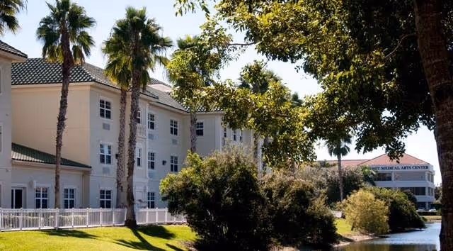 Exterior view of a multi-story building with white walls and green roof tiles, surrounded by palm trees and other greenery. A white fence runs along the front of the building, and there is a body of water with bushes and trees nearby. Another building labeled 'Medical Arts Center' is visible in the background.