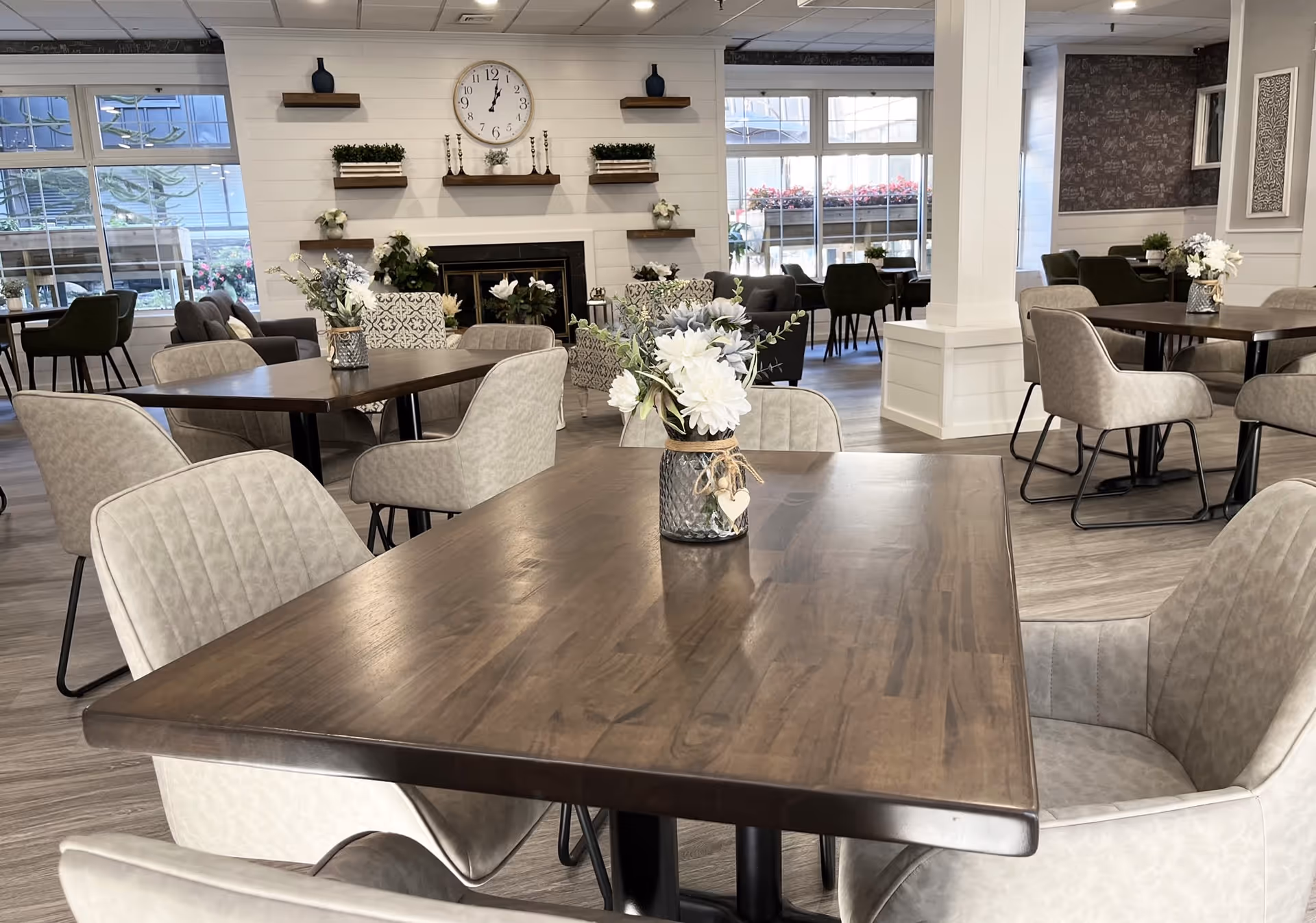 A bright and spacious dining area with multiple wooden tables and cushioned chairs. Each table has a decorative vase with white flowers. The room features large windows letting in natural light, a fireplace with shelves above it holding plants and decorative items, and a large clock on the wall. The flooring is light wood, and the overall decor is modern and inviting.