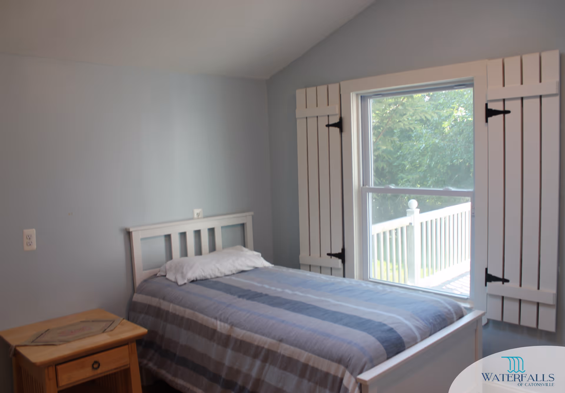 Small bedroom with a single bed against a pale blue wall, a window with white shutters overlooking a railing, and a wooden bedside table.