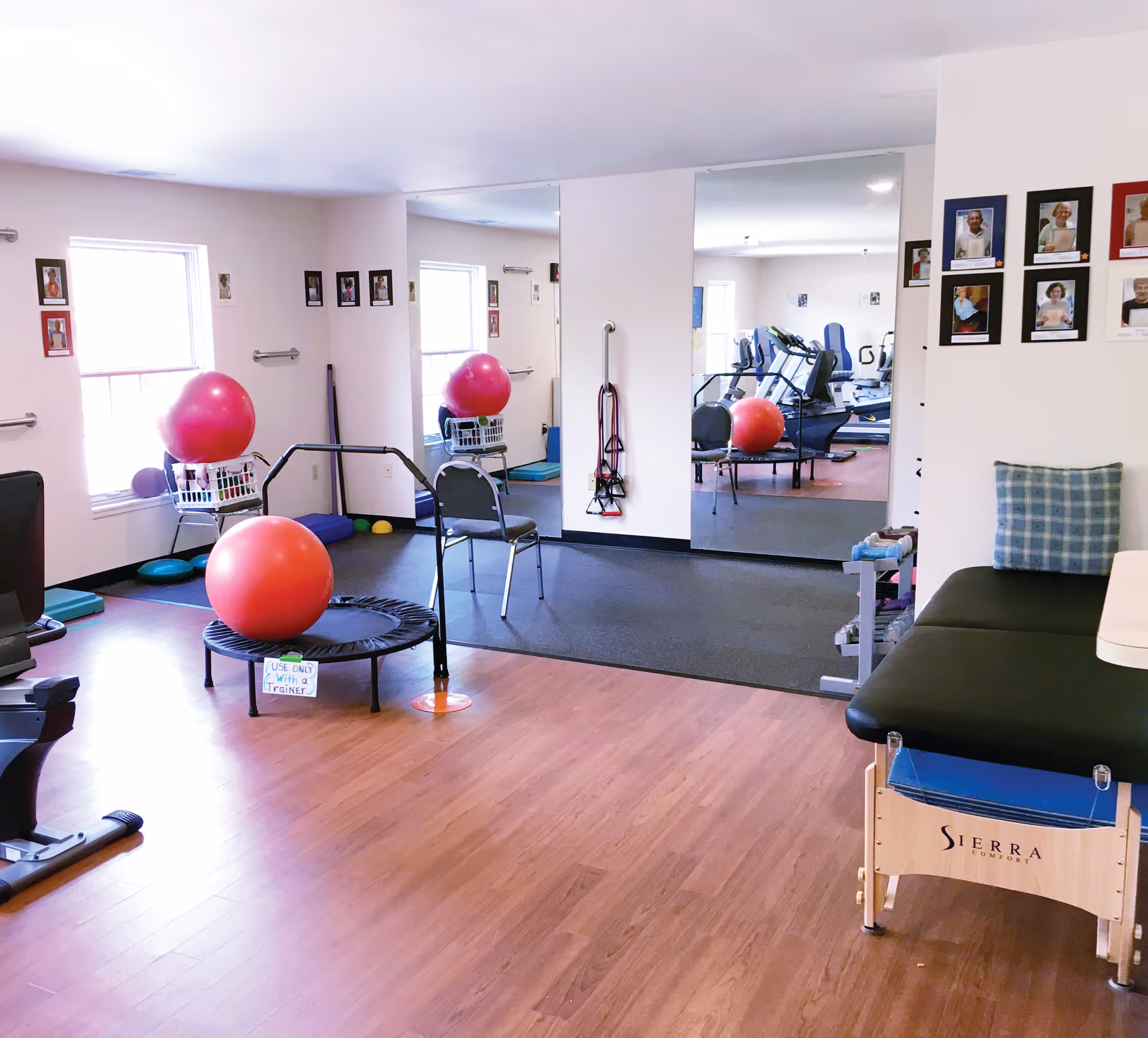 A senior living facility exercise room with wooden floors and light-colored walls. The room contains exercise equipment including a mini trampoline with a large red exercise ball on it, a chair, resistance bands hanging on the wall, and a therapy table with a blue checkered pillow. There are large mirrors on one wall reflecting more exercise equipment such as treadmills and another red exercise ball. Several framed photos of residents or staff are displayed on the walls.