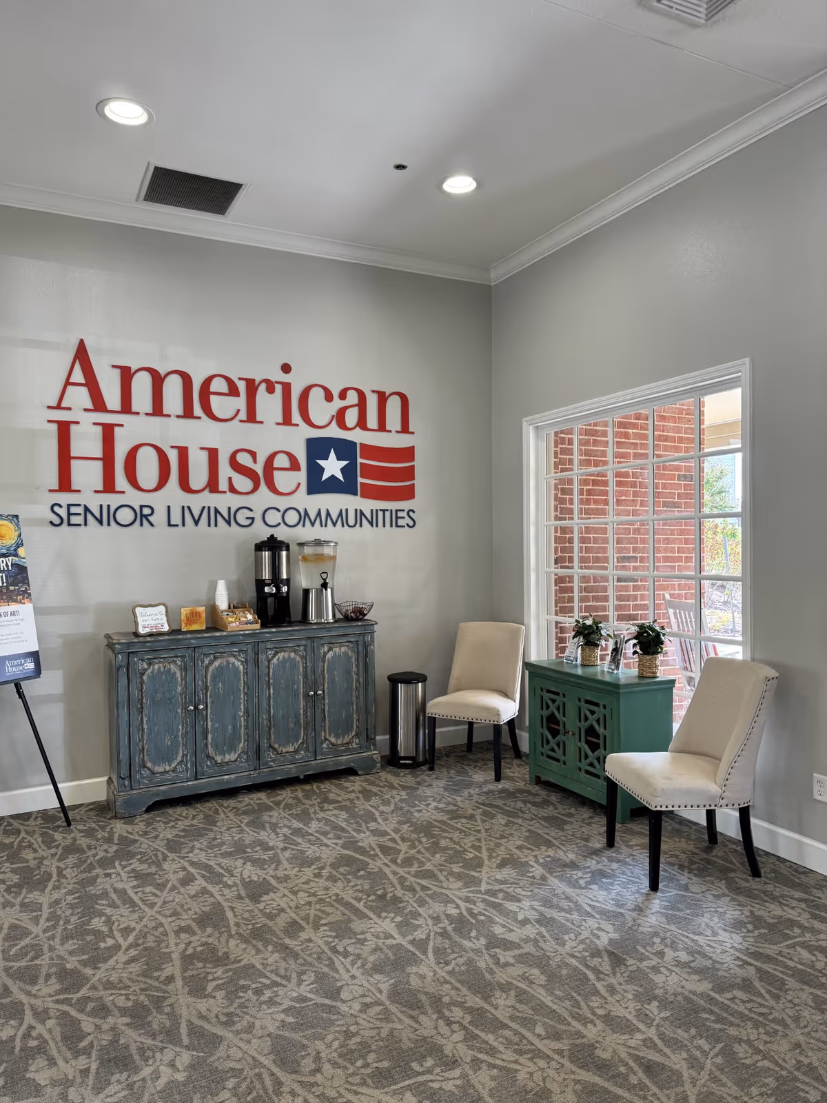 Lobby seating area with an "American House Senior Living Communities" logo on the wall, a beverage station on a distressed cabinet, two chairs and a small table by a window.