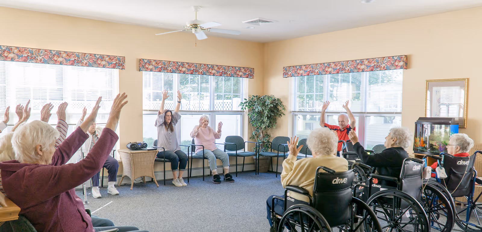 A group of elderly people, some in wheelchairs, and a caregiver sitting in a circle in a bright room with large windows, raising their hands as part of a group activity or exercise. The room has beige walls, floral valances on the windows, a ceiling fan, a potted plant, and a fish tank.