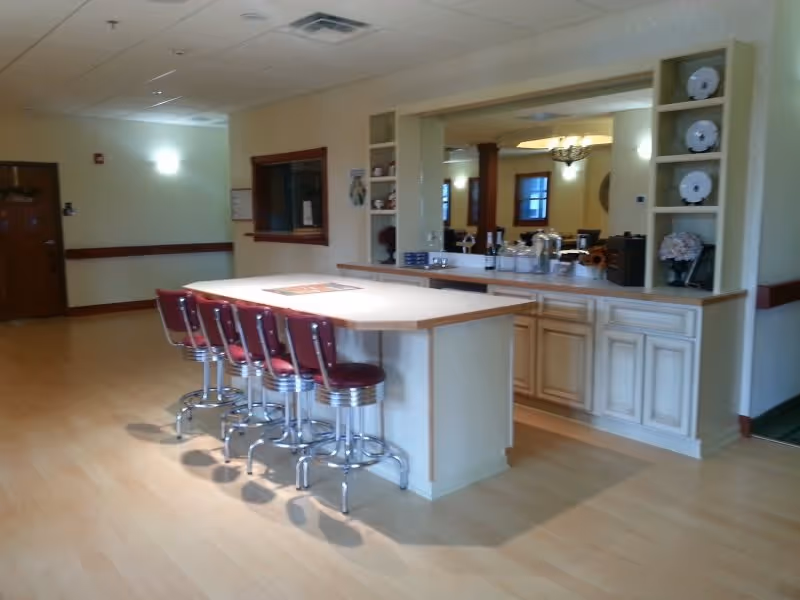 Interior view of a senior living facility showing a kitchen island with five red cushioned bar stools. The island has a light wood countertop and cream-colored cabinets underneath. Behind the island, there is a counter with decorative plates on shelves and various items including a coffee maker and glassware. The room has light-colored walls and wood flooring, with a window and a door visible in the background.