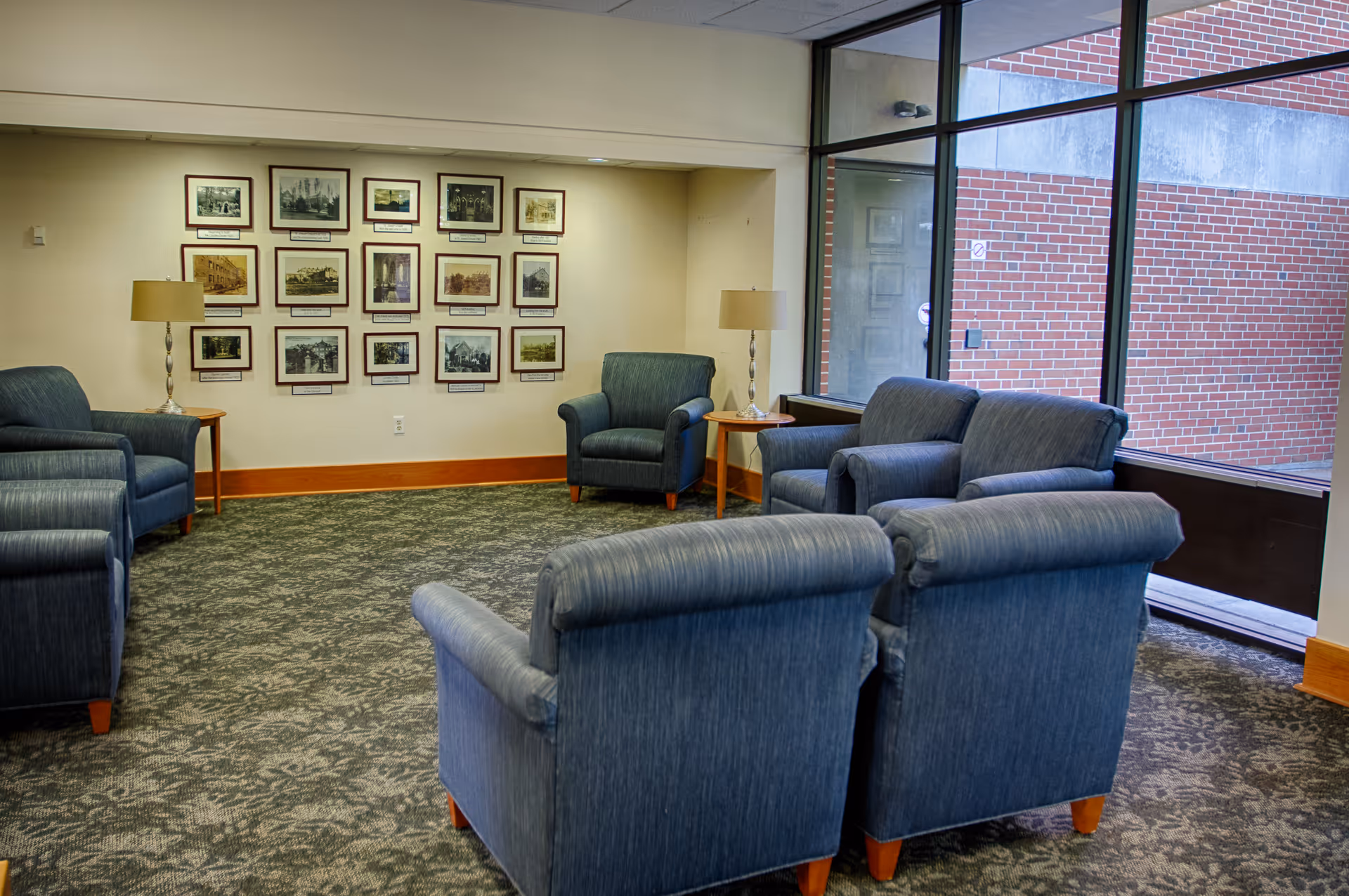 A senior-living lounge with several blue upholstered armchairs arranged around side tables, framed photos on a wall, and large windows showing a brick exterior.