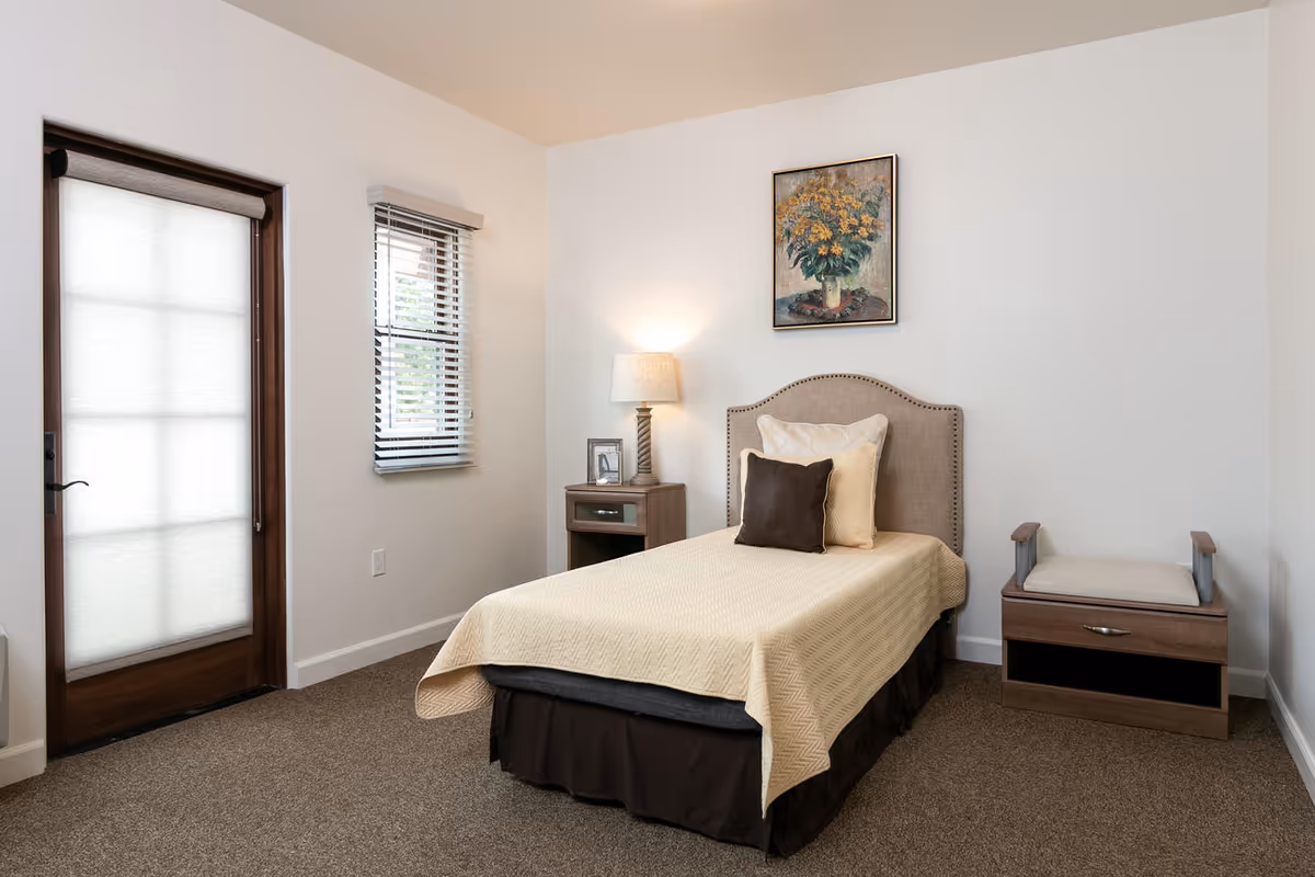 Simple single-occupancy bedroom with a bed, nightstand and lamp, wall art, and a frosted-glass door and window.
