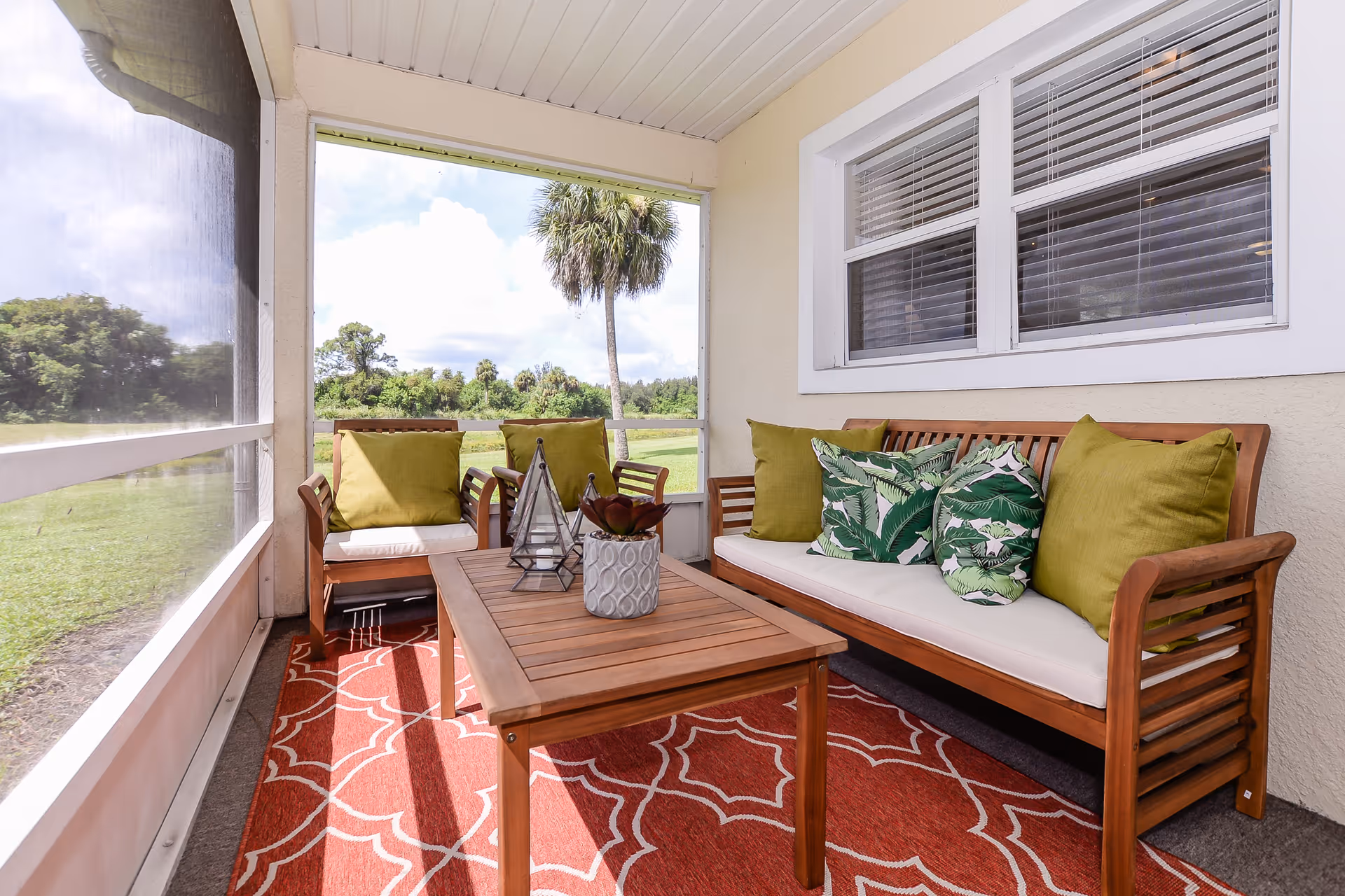Screened porch with wooden sofa and chairs, green and patterned pillows, a wooden coffee table on a red patterned rug, and a grassy yard with a palm tree visible outside.