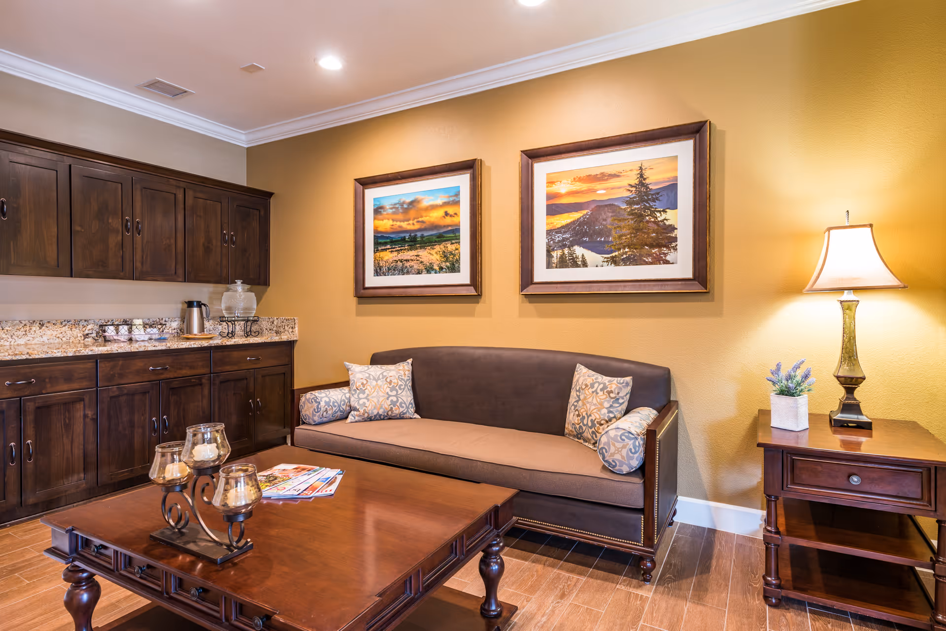 A cozy interior room with a brown sofa adorned with patterned pillows, a wooden coffee table with candle holders and magazines, dark wooden cabinets with a granite countertop holding a coffee pot and cups, two framed landscape paintings on a yellow wall, and a wooden side table with a lamp and a small potted plant.