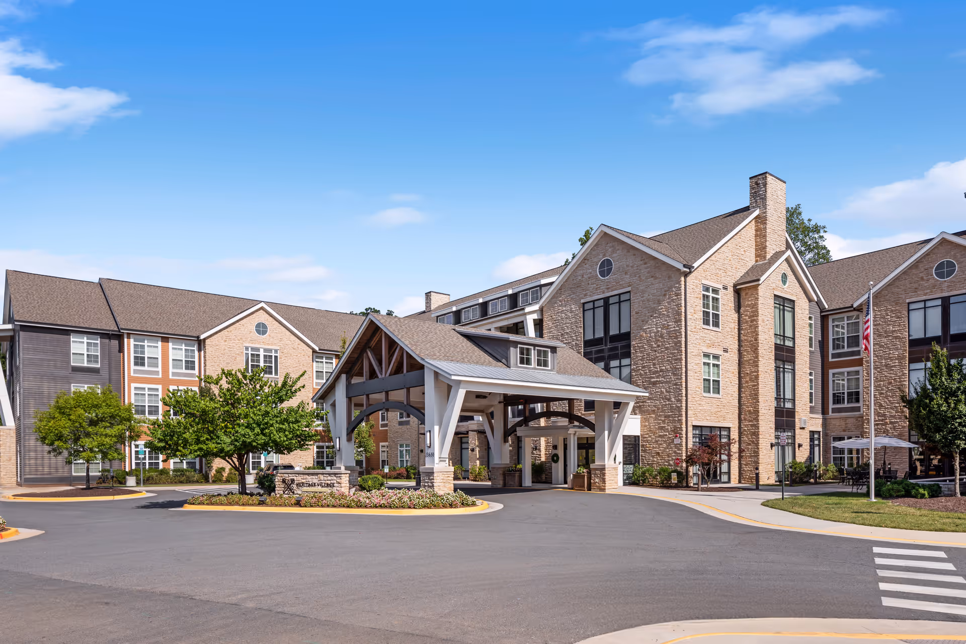 Exterior view of Heritage Village Assisted Living and Memory Care facility showing a large building with multiple windows, a covered entrance with wooden beams, landscaped greenery, and a clear blue sky.