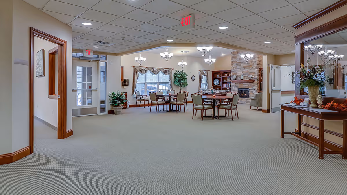 Interior view of a senior living facility common area with carpeted floors, multiple round tables with chairs, a stone fireplace, large windows with curtains, plants, and ceiling chandeliers providing lighting.