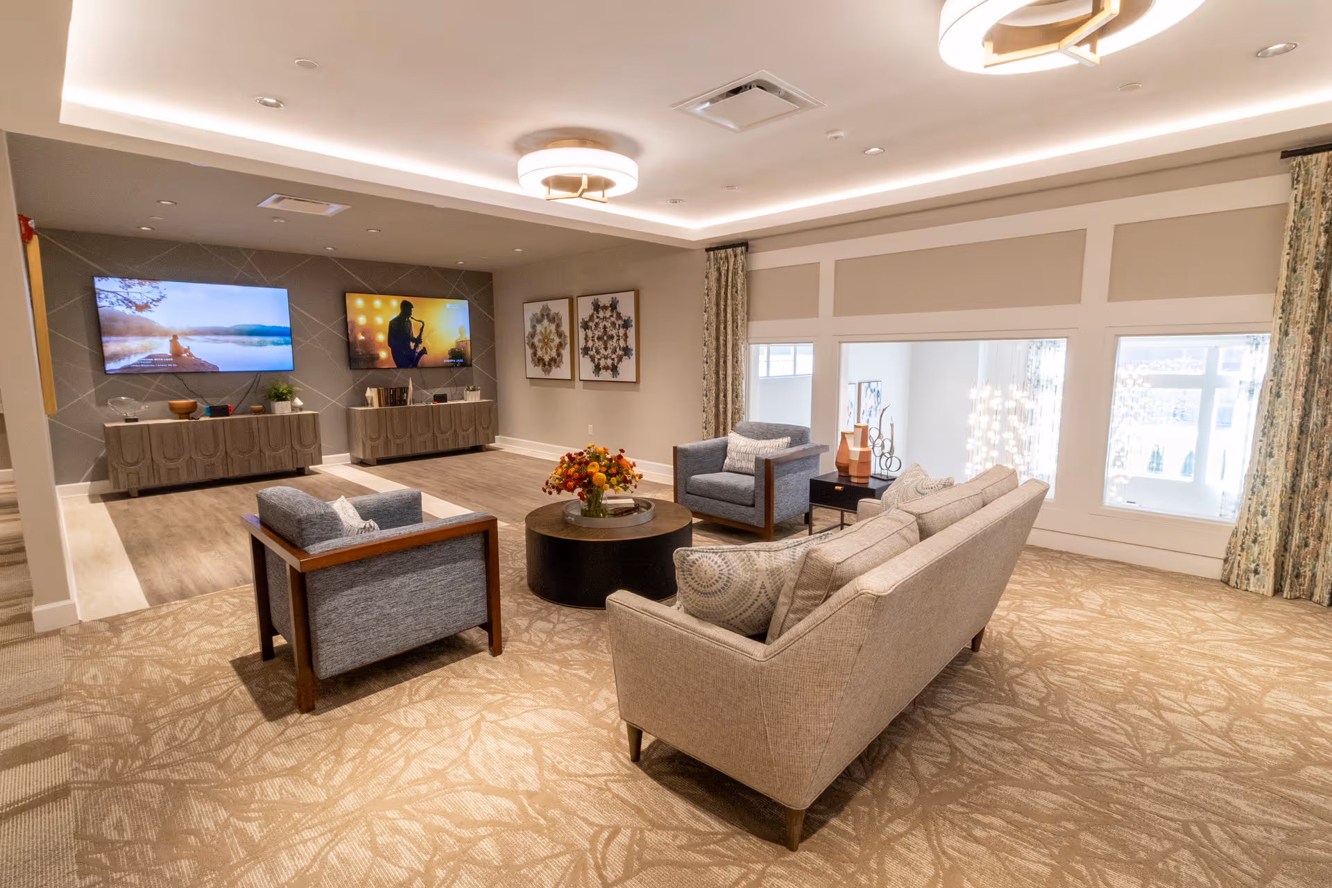 Spacious communal living room with sofas and armchairs arranged around a central coffee table, two wall-mounted TVs, and decorative artwork.