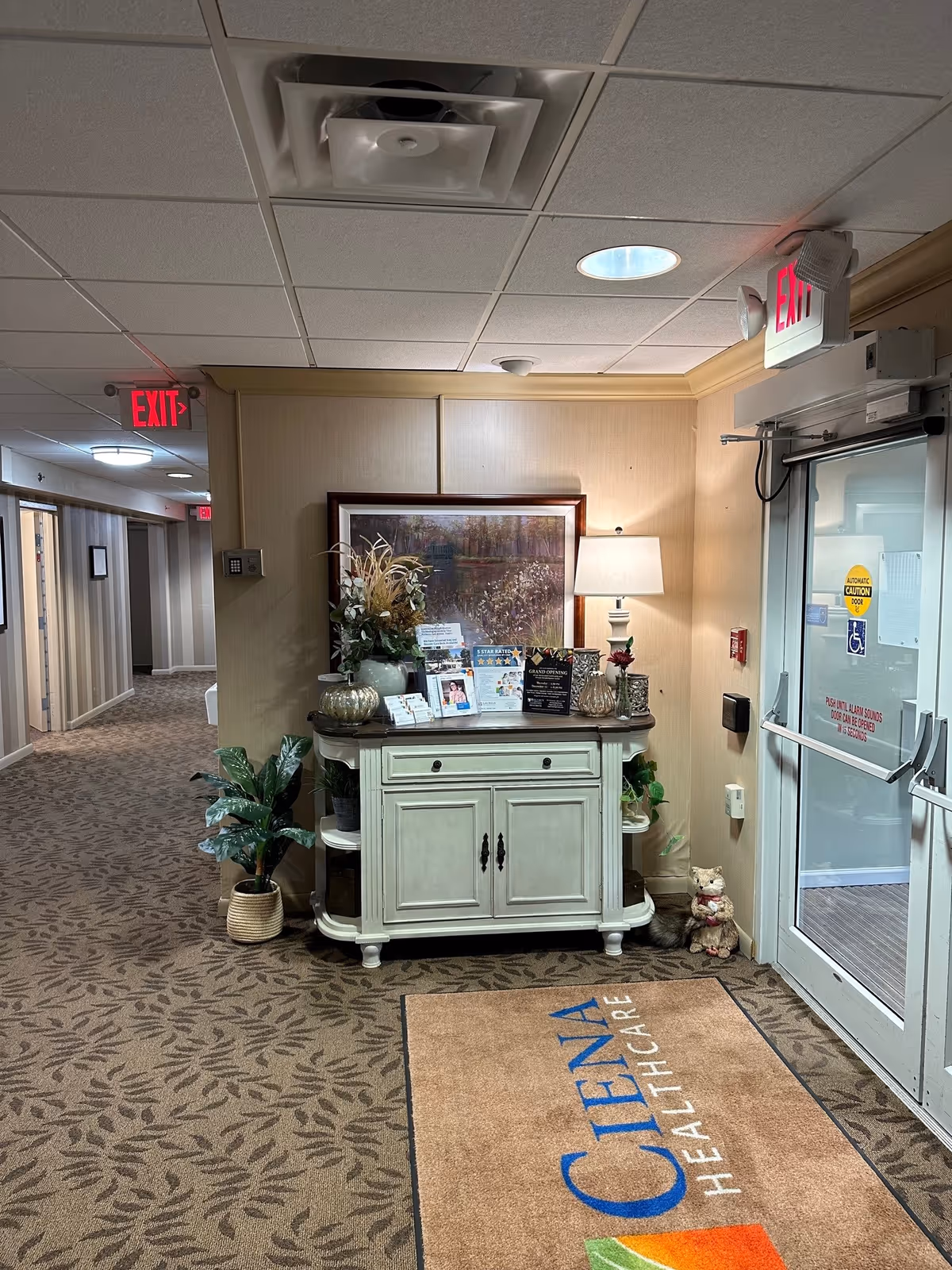 Interior entry hallway with a decorative console table topped with a lamp and artwork beside a glass exit door and a branded floor mat.