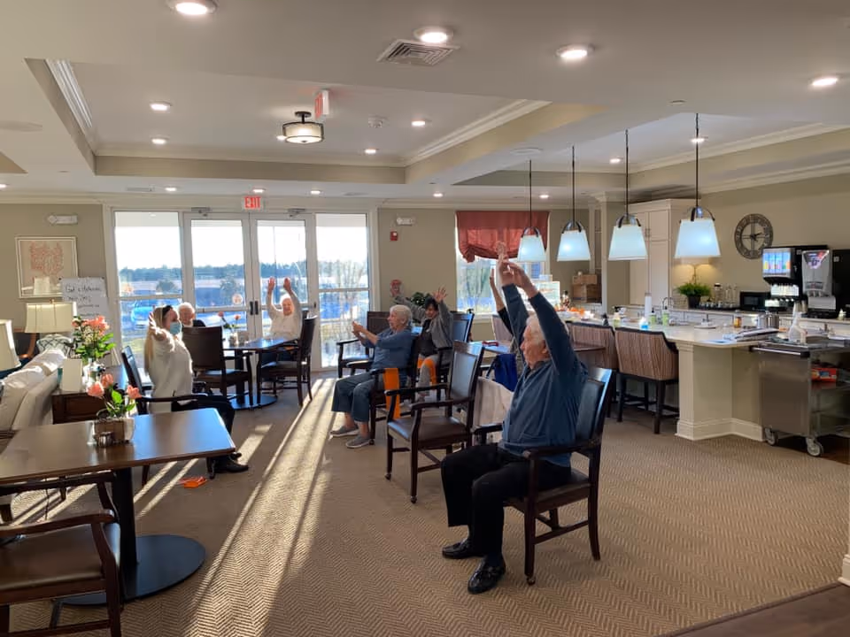 A group of elderly people seated in chairs in a well-lit common area of a senior living facility, participating in a seated exercise or stretching activity with their arms raised. The room features large windows letting in natural light, a kitchen area with pendant lights, tables, chairs, and a beverage station.