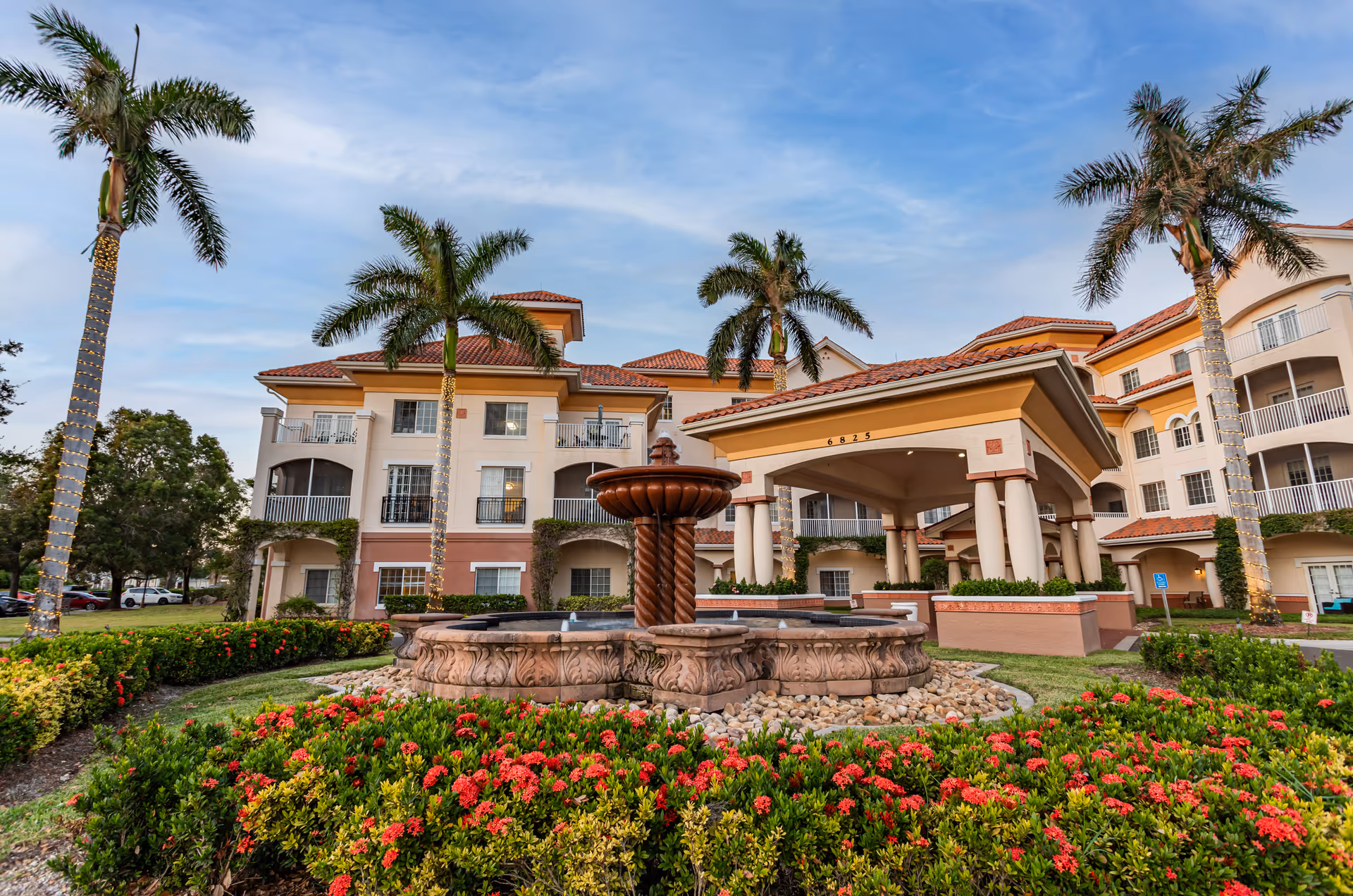 Exterior view of Terracina Senior Living facility featuring a multi-story building with balconies, a covered entrance with columns, a decorative fountain in the foreground, palm trees wrapped with lights, and landscaped gardens with red flowers under a partly cloudy sky.