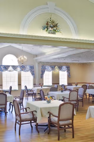 Bright dining room with multiple small tables covered in white tablecloths, floral centerpieces, cushioned wooden chairs, chandeliers, and arched windows with valances.