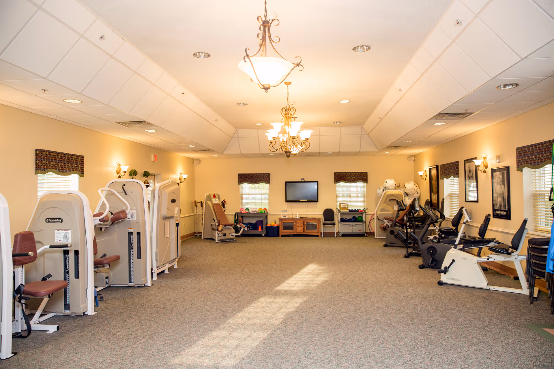Spacious senior fitness room with exercise machines, recumbent bikes, a wall-mounted TV and chandeliers.