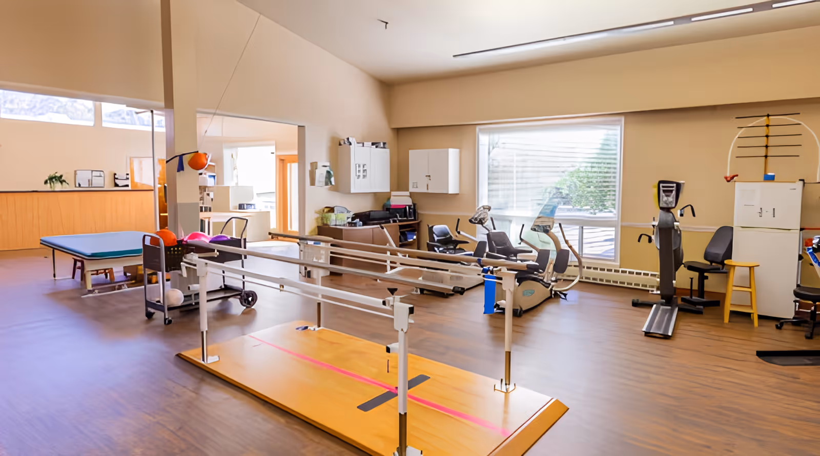 A spacious rehabilitation room in a nursing center featuring parallel bars for walking exercises, stationary exercise bikes, a therapy table, and various chairs. The room has large windows allowing natural light and light-colored walls with wooden flooring.