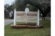 Outdoor image of a white sign for Trinity Mission Health & Rehab of Granbury located on a grassy area with trees and a road in the background.
