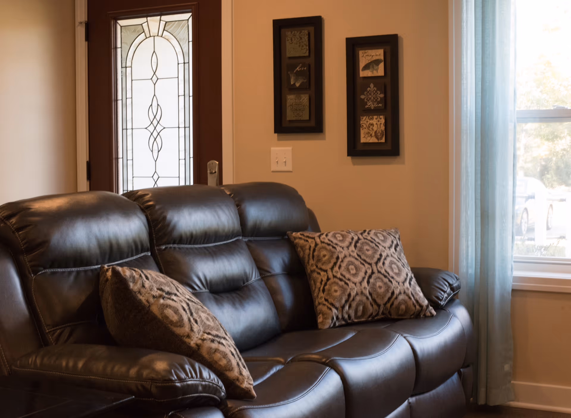 A cozy living room area featuring a dark brown leather sofa with two patterned throw pillows. Behind the sofa is a decorative glass front door and two framed wall art pieces. To the right, there is a window with light blue curtains letting in natural light.