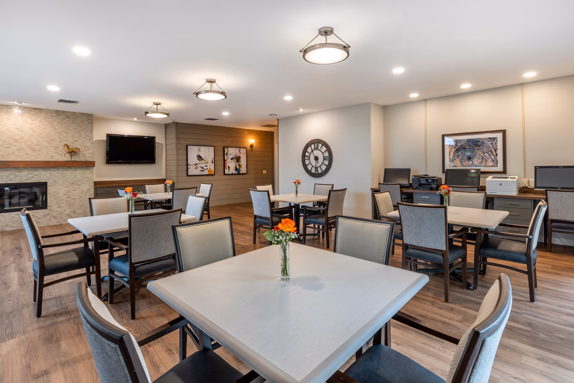 Bright communal dining room with multiple tables and chairs, small vases of flowers, a wall clock, TV, and a fireplace.