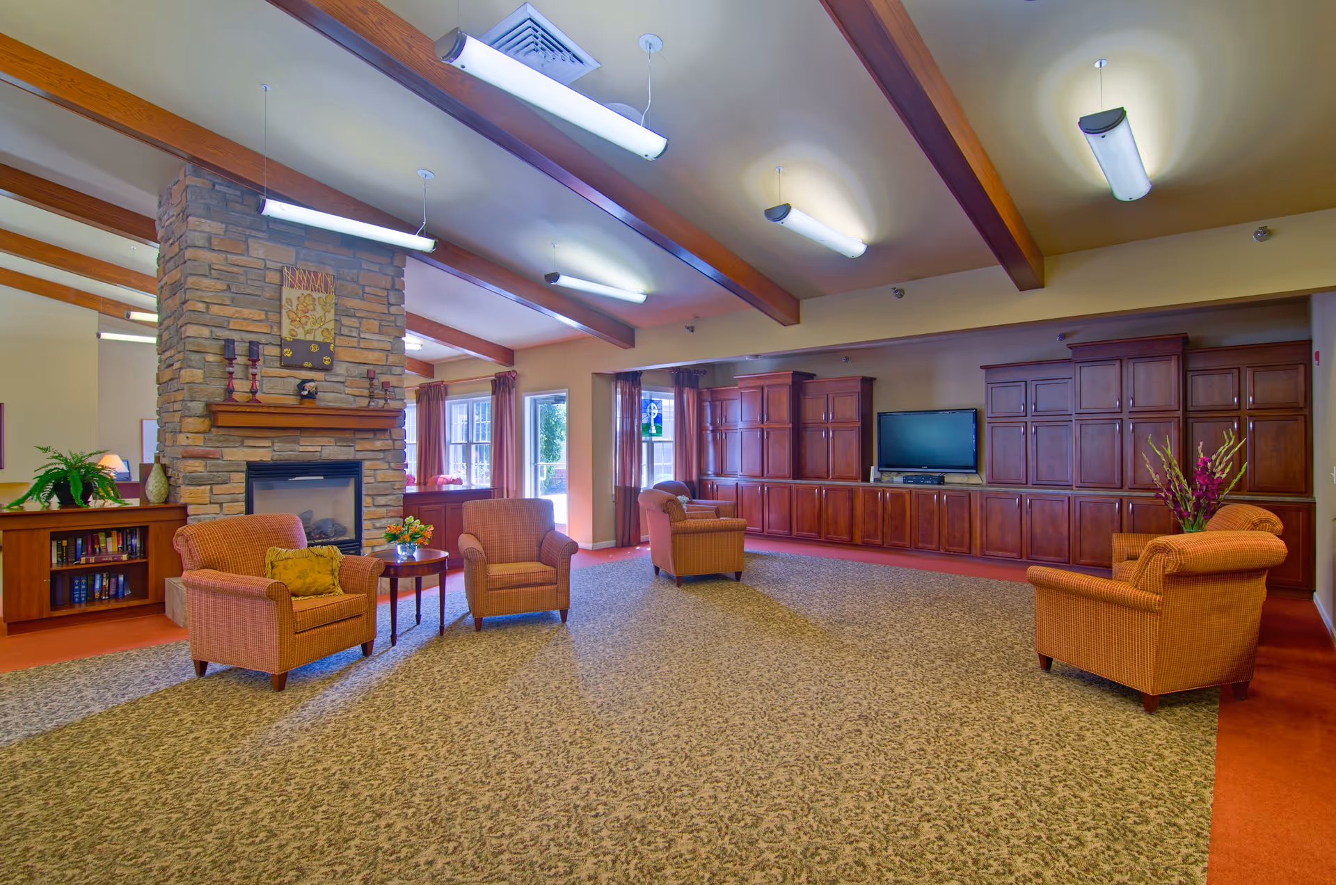 A spacious living room area in an assisted living facility featuring four cushioned armchairs arranged around a small wooden table with a flower vase. The room has a stone fireplace with decorative items on the mantel, large wooden cabinets along one wall with a flat-screen TV, and large windows with curtains letting in natural light. The ceiling has exposed wooden beams and fluorescent lighting fixtures.