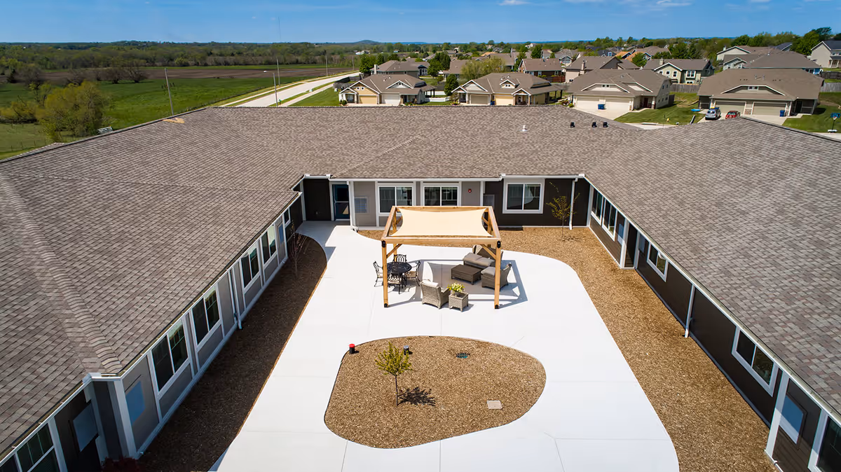 Aerial view of a single-story assisted living facility arranged in a U-shape around a central courtyard. The courtyard features a shaded seating area with chairs and a small table, surrounded by a concrete walkway and landscaped with mulch and small trees. Residential houses and green fields are visible in the background under a clear blue sky.