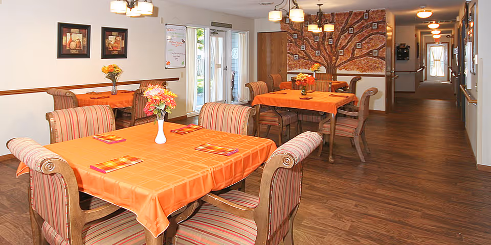 A dining area in a senior living facility with several tables covered in orange tablecloths, each adorned with a vase of flowers. The room has wooden flooring, striped cushioned chairs, framed artwork on the walls, and a large decorative tree mural with photos on the far wall. A hallway extends from the dining area, and glass doors lead outside.