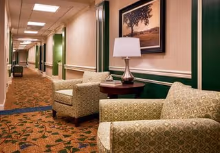 A long hallway in a senior living facility with patterned carpet and green and beige walls. Two cushioned armchairs and a small round table with a lamp and a framed picture are placed along the wall.
