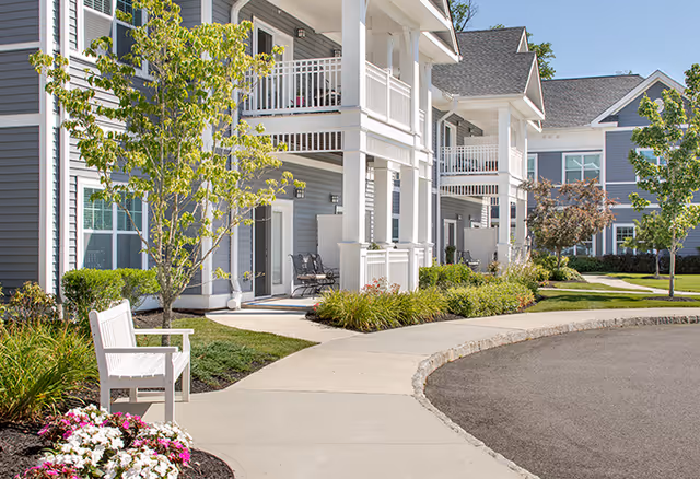 Exterior front of a two-story senior living building with white balconies, a landscaped walkway, and a bench.