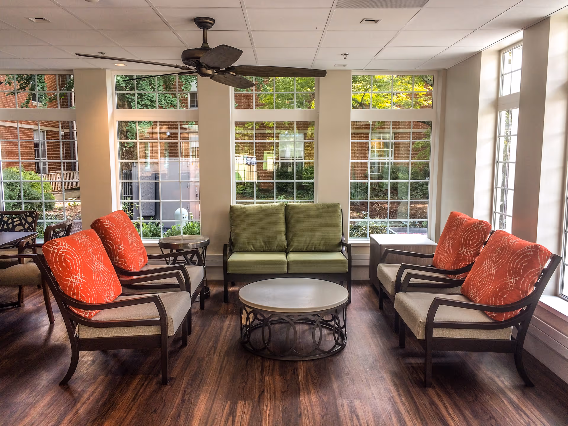 A bright and cozy sitting area with large windows showing a garden outside. The room features four armchairs with orange patterned cushions, a green cushioned loveseat, a round coffee table, and side tables. The floor is wooden, and a ceiling fan is mounted above.
