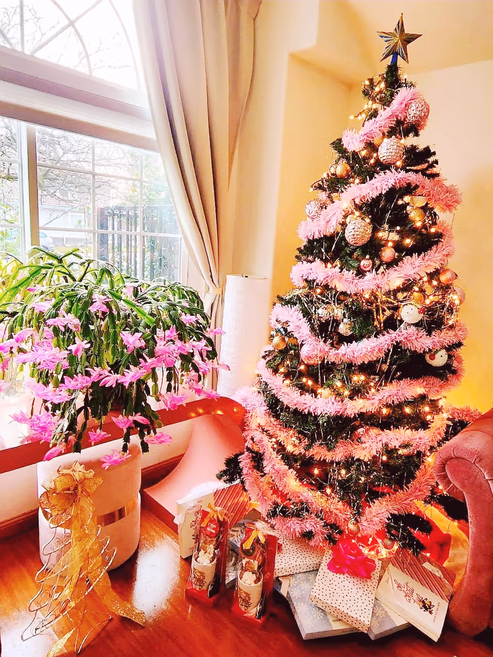 A decorated Christmas tree with pink garlands, gold and white ornaments, and a star topper stands next to a large window with beige curtains. There are wrapped gifts under the tree and a potted plant with pink flowers beside it. A pink armchair is partially visible on the right side.