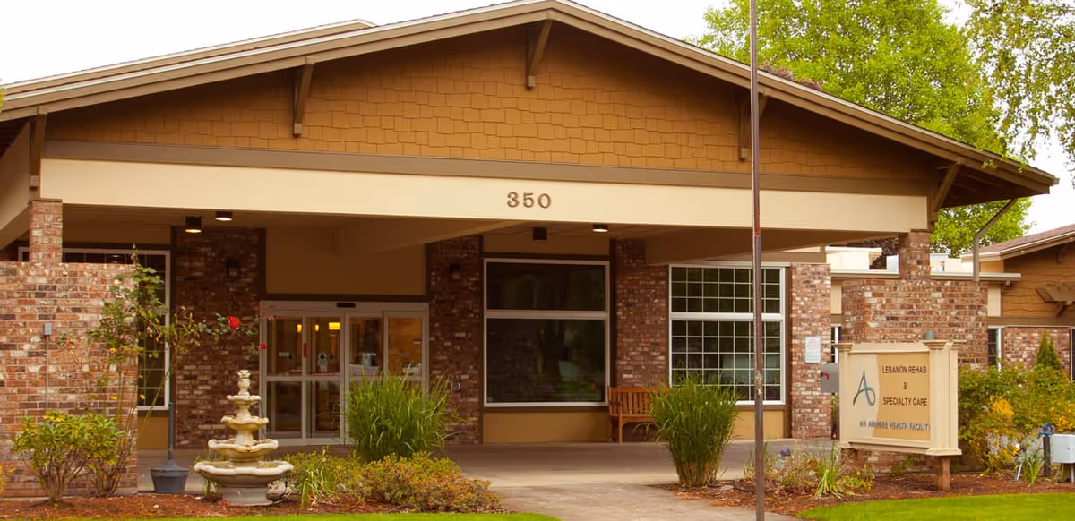 Covered brick front entrance of a senior living facility with the number '350' above the doorway, a small fountain, bench, sign, and landscaped greenery.
