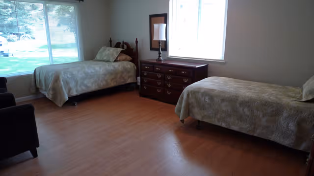 A simple bedroom with two single beds covered in matching beige patterned bedspreads. Between the beds is a wooden dresser with a lamp and a mirror above it. There are two large windows letting in natural light, and the floor is wooden.