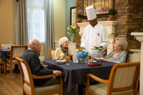 A chef serving food to three elderly people seated around a dining table in a cozy room with a stone fireplace and framed artwork on the wall.
