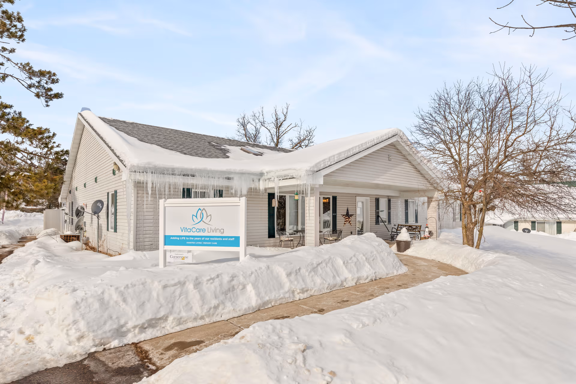 Exterior view of VitaCare Living Spooner facility in winter, showing a single-story building with a snow-covered roof and icicles hanging from the eaves. Snow blankets the ground and surrounds a cleared walkway leading to the entrance. A sign with the VitaCare Living logo and tagline is visible in front of the building. Leafless trees and a clear sky are in the background.