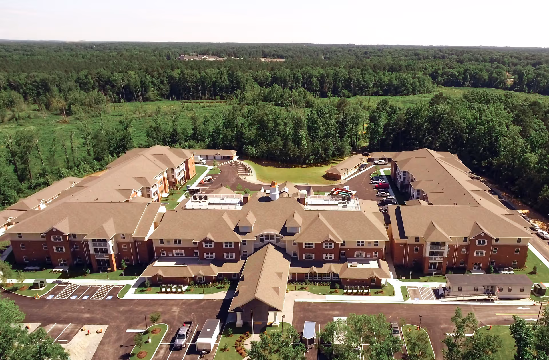 Aerial view of Tuckahoe Pines Retirement Community showing a large, multi-wing brick building with a brown roof surrounded by parking lots and green trees in the background.