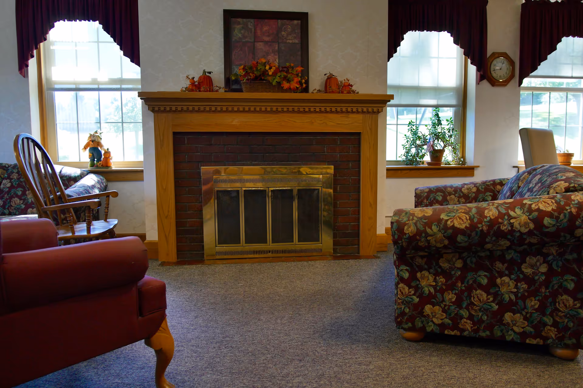 A cozy common room with a central brick fireplace, mantel decorations, windows, and floral upholstered chairs.