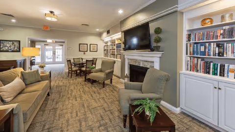 A cozy living room area in Sycamore Manor featuring a beige sofa with cushions, two green armchairs, a wooden coffee table with a potted plant, a fireplace with a mounted flat-screen TV above it, built-in bookshelves filled with books, and a dining table with chairs in the background near a glass door exit.