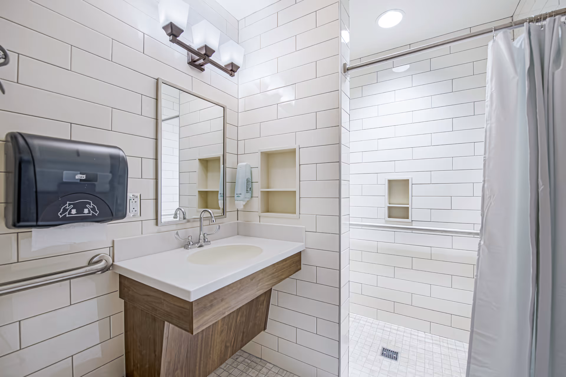 A clean, modern bathroom with white tiled walls and floor. The image shows a sink with a large mirror above it, a soap dispenser mounted on the wall, a paper towel dispenser, and a walk-in shower with a curtain and built-in shelves.