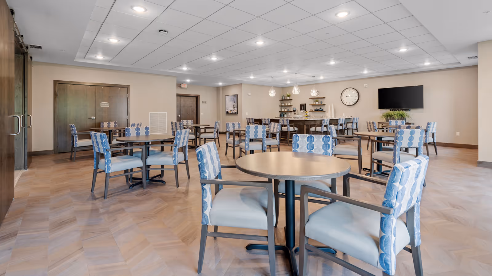 A spacious dining room with multiple round tables and chairs featuring blue patterned upholstery. The room has a light wood floor, beige walls, recessed ceiling lights, a wall clock, a flat-screen TV, and shelves with decorative items and plants.