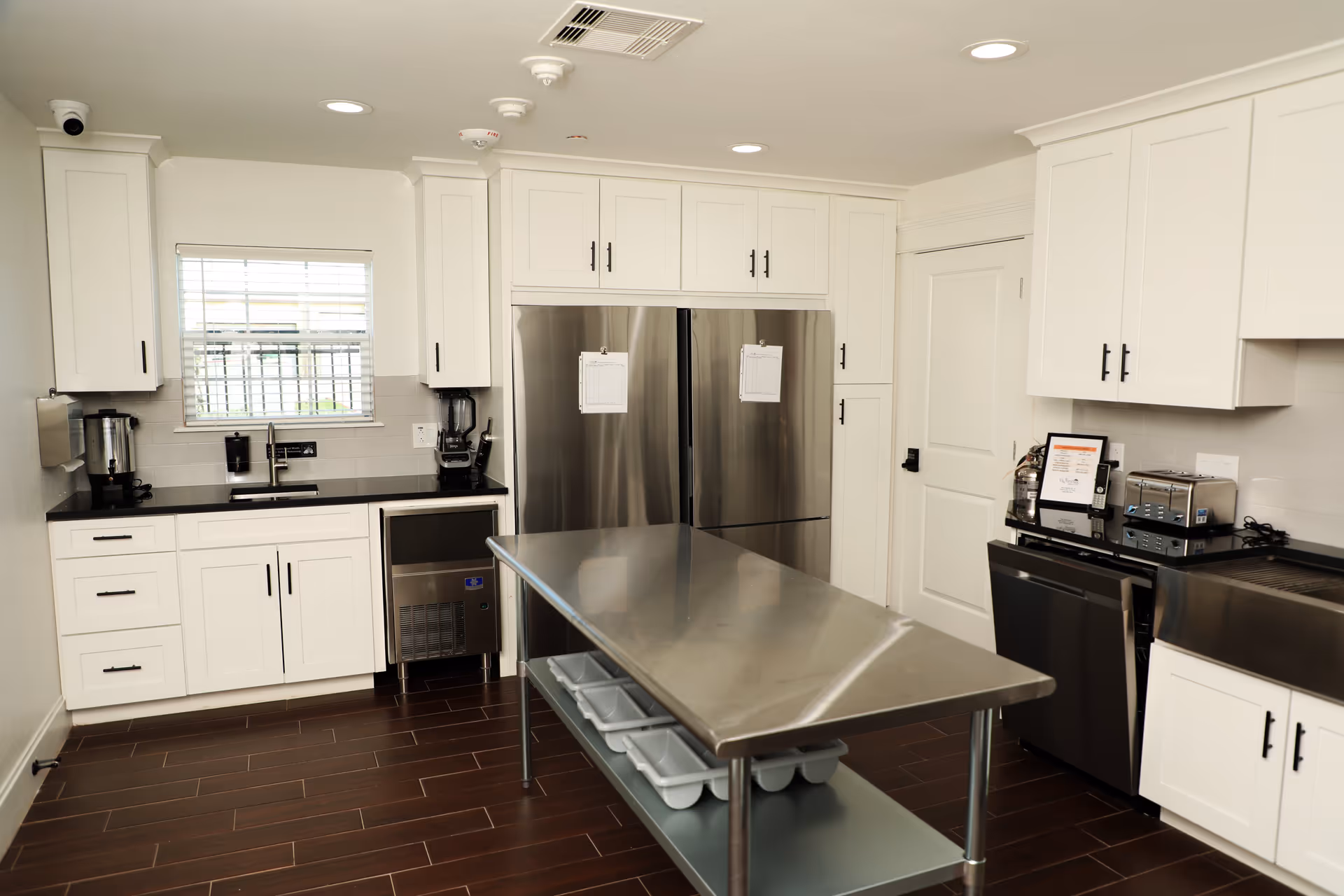 A clean, modern kitchen with white cabinets, stainless steel appliances including two large refrigerators, a dishwasher, and a toaster. There is a stainless steel island table in the center with trays underneath. The kitchen has dark wood flooring and a small window with blinds above the sink.