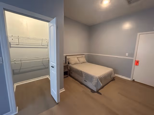 A simple bedroom with a single bed covered in beige bedding, a small nightstand beside it, light gray walls with white trim, and a door leading to a walk-in closet with wire shelving. The floor is a light wood tone.